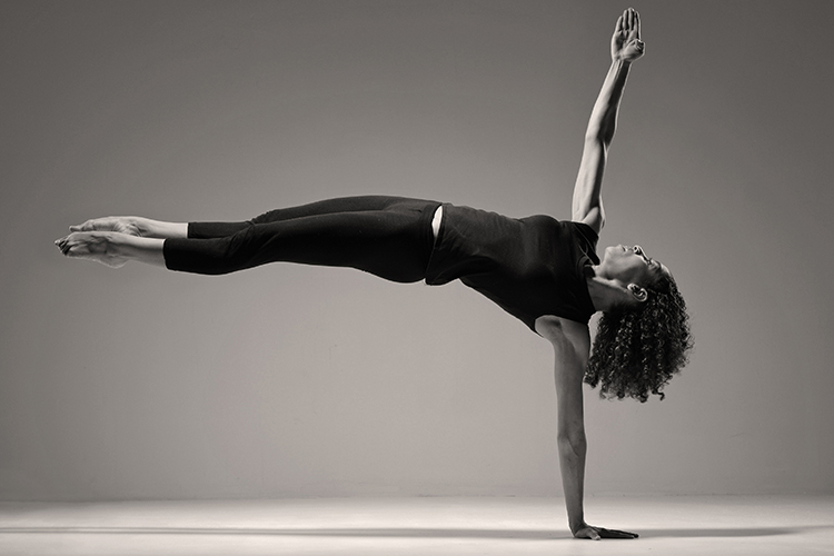 A dancer performs a handstand with her body perfectly parallel to the floor while her other arm extended upward perpendicularly. She has long curly hair and is wearing black activewear.