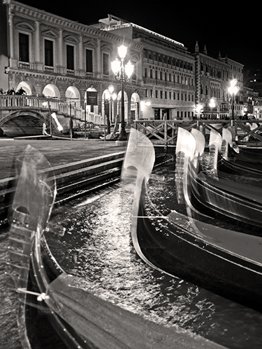 Black and white photo of gondolas lined up on a canal at night. Street lamps illuminate the historic buildings and arched bridge in the background, creating reflections on the water.